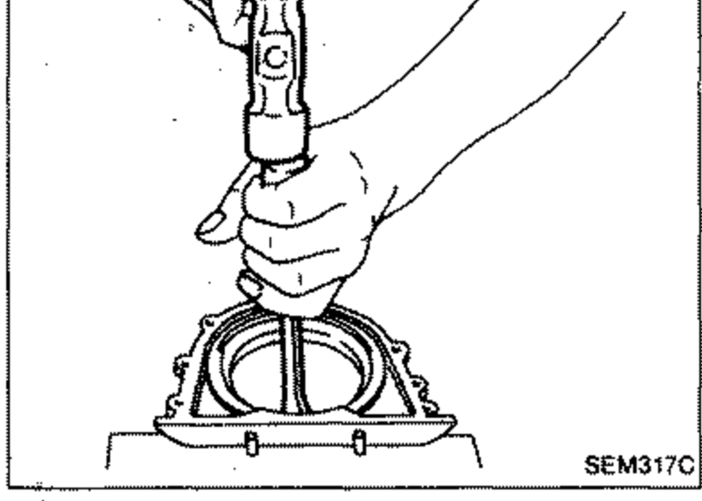 Illustration of removing the seal from the rear oil seal retainer using a tool. A hand presses a tool down onto the retainer mounted on a fixture.