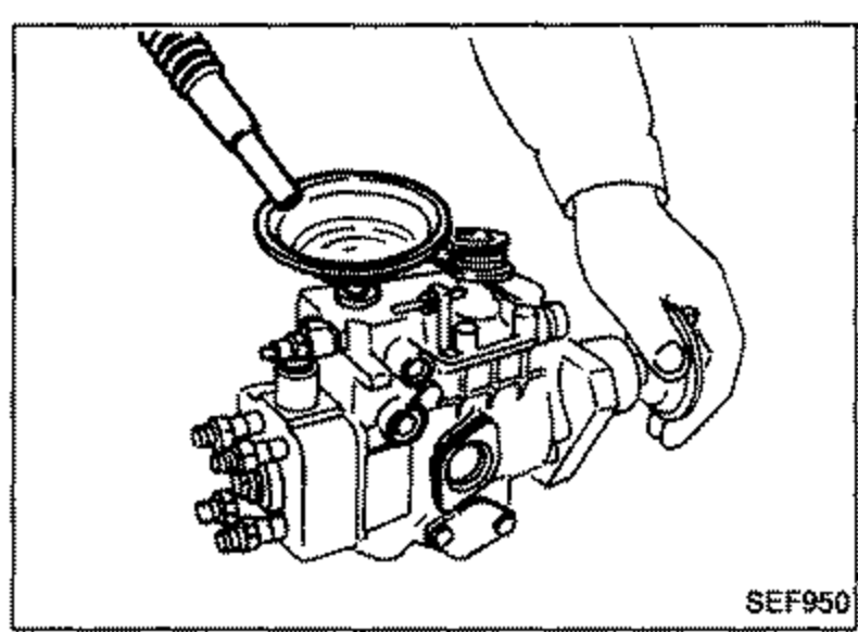Test oil being poured into the fuel injection pump from a spouted container held by a hand, viewed from the top of the pump.
